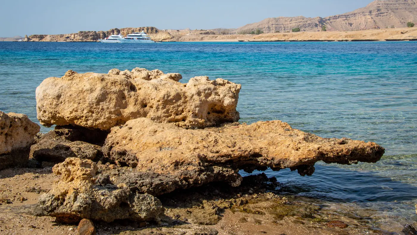 A tranquil beach scene featuring rocky shores, clear blue waters, and distant boats under a summer sky.