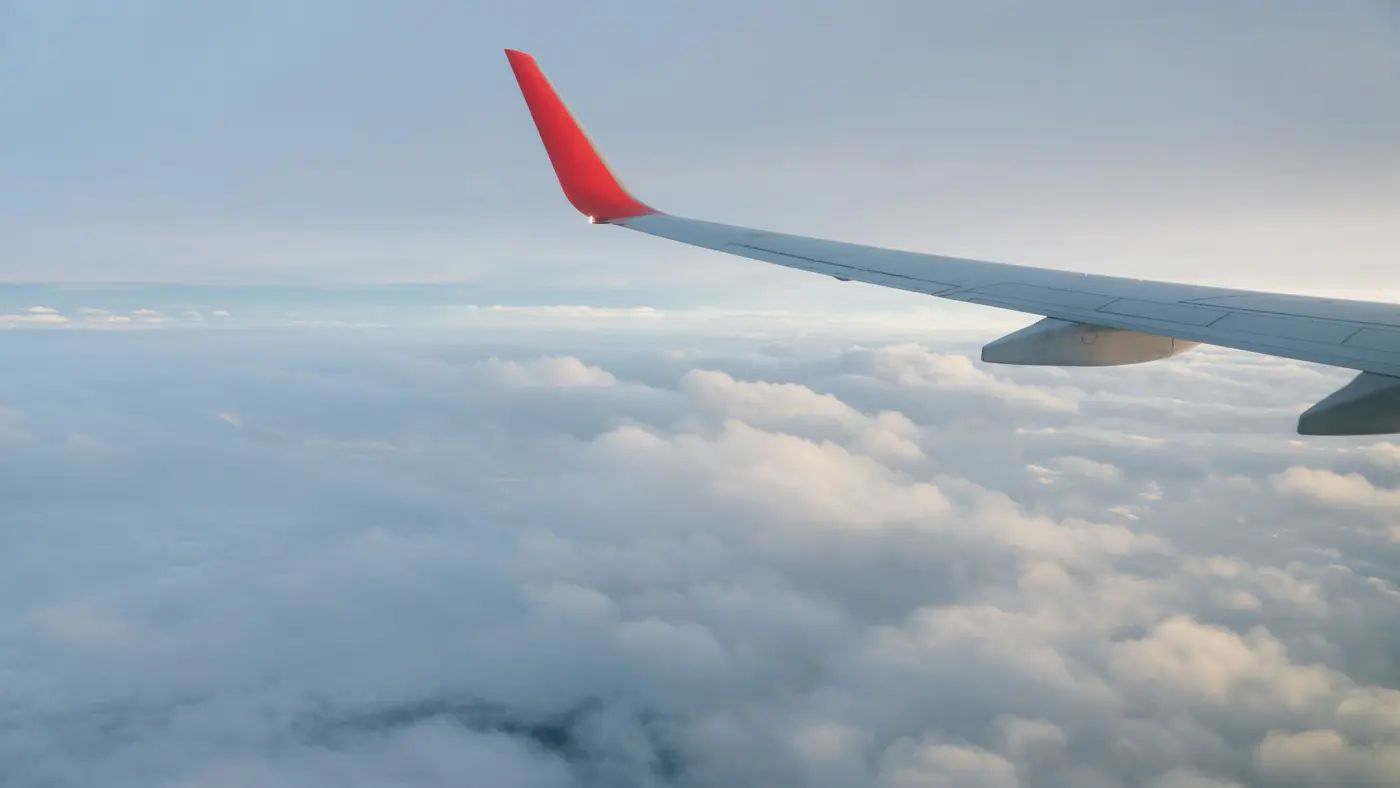Through window of red and white wing of aircraft flying over fluffy clouds in endless blue sky