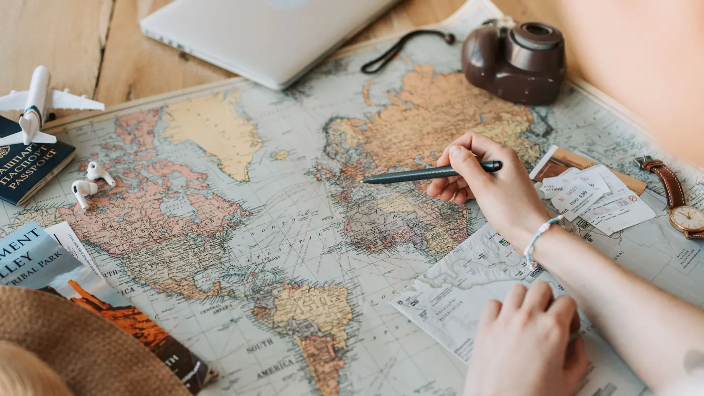 Hands marking destinations on a world map surrounded by travel essentials like passport and camera.