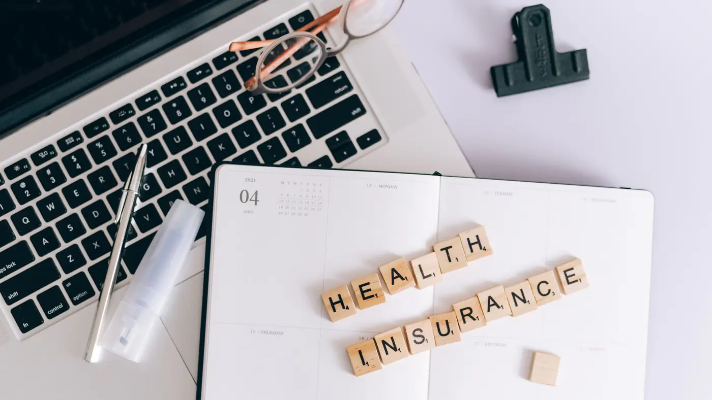 Scrabble tiles spelling health insurance on a planner next to a laptop.