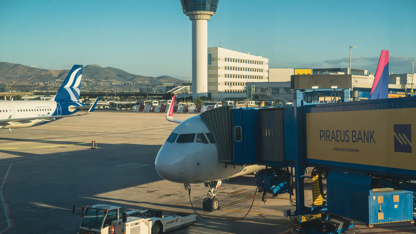 An airplane at Athens International Airport gate during a sunny day, featuring a jet bridge and terminal view.