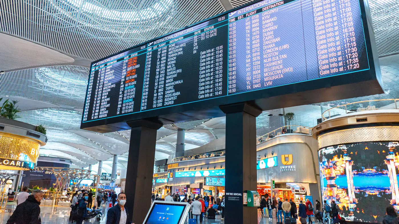 Passengers walk through Istanbul Airport's departure area, surrounded by stores and flight information displays.
