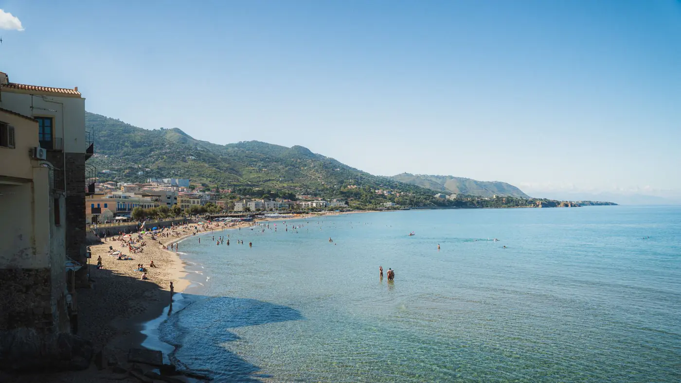 Idyllic coastal scene with a beach, mountains, and clear blue sea under a sunny sky.