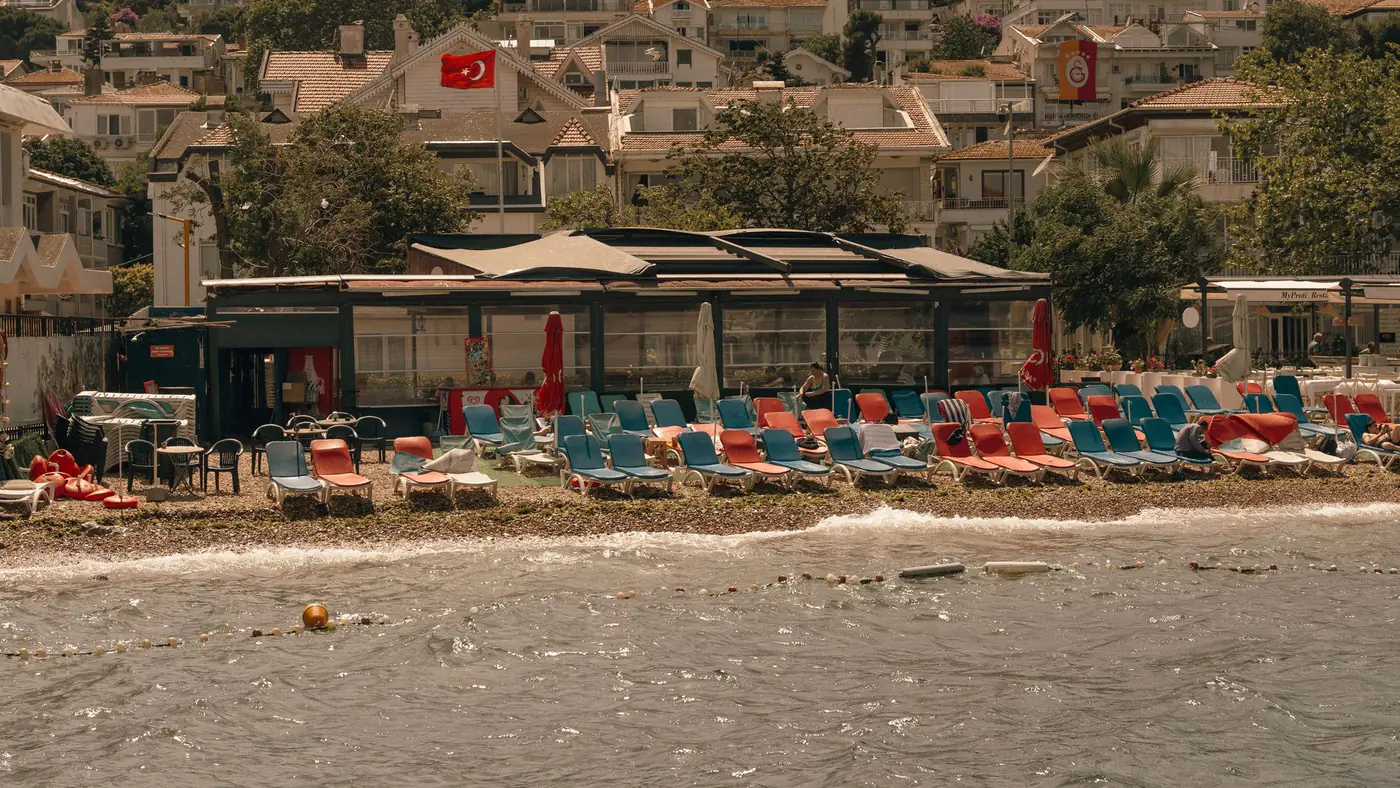 Empty beach with lounge chairs and Turkish flags against a backdrop of coastal homes.