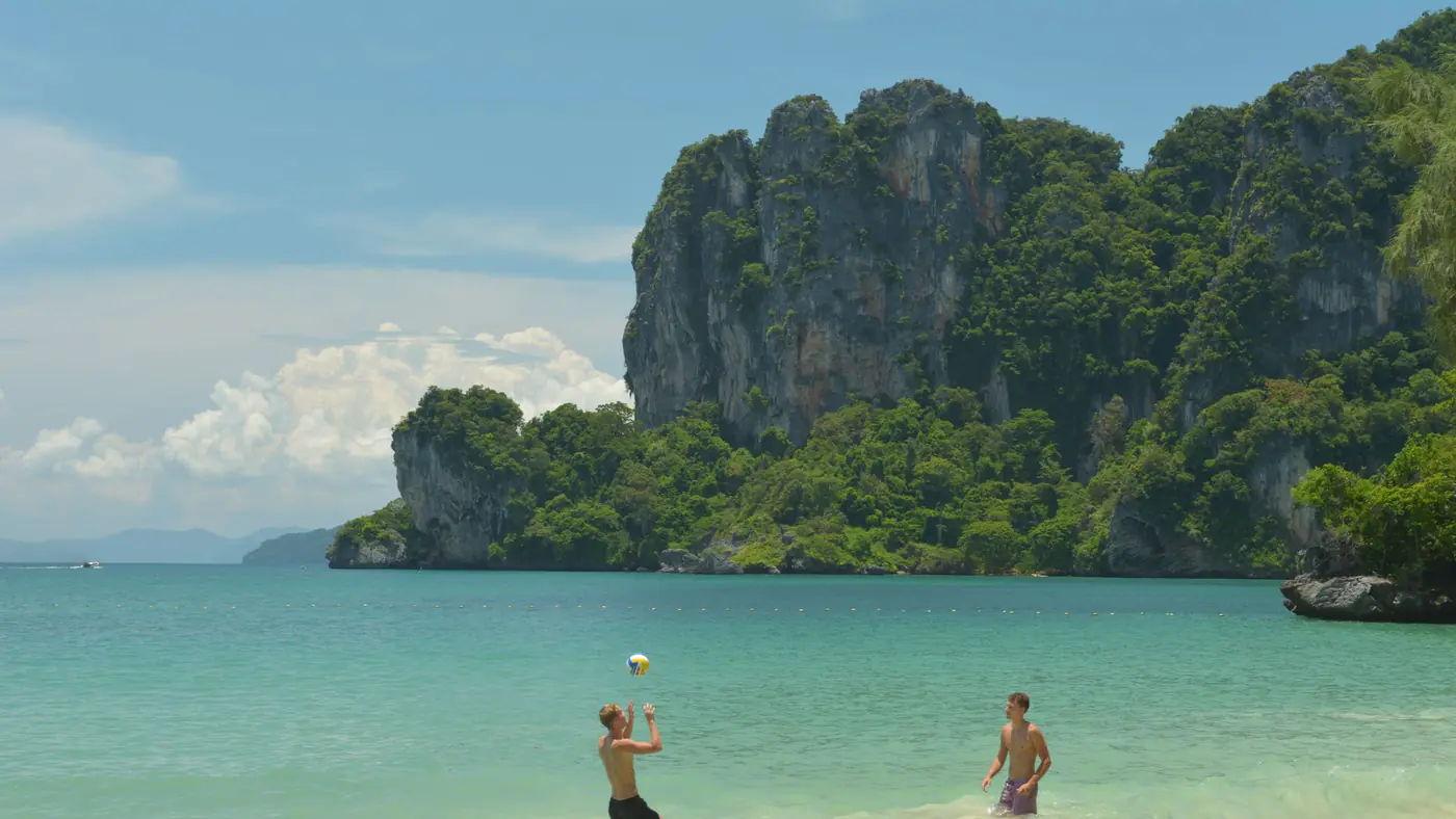Two men enjoy a game of beach volleyball on Ao Nang's sandy shores with stunning limestone cliffs in the background.