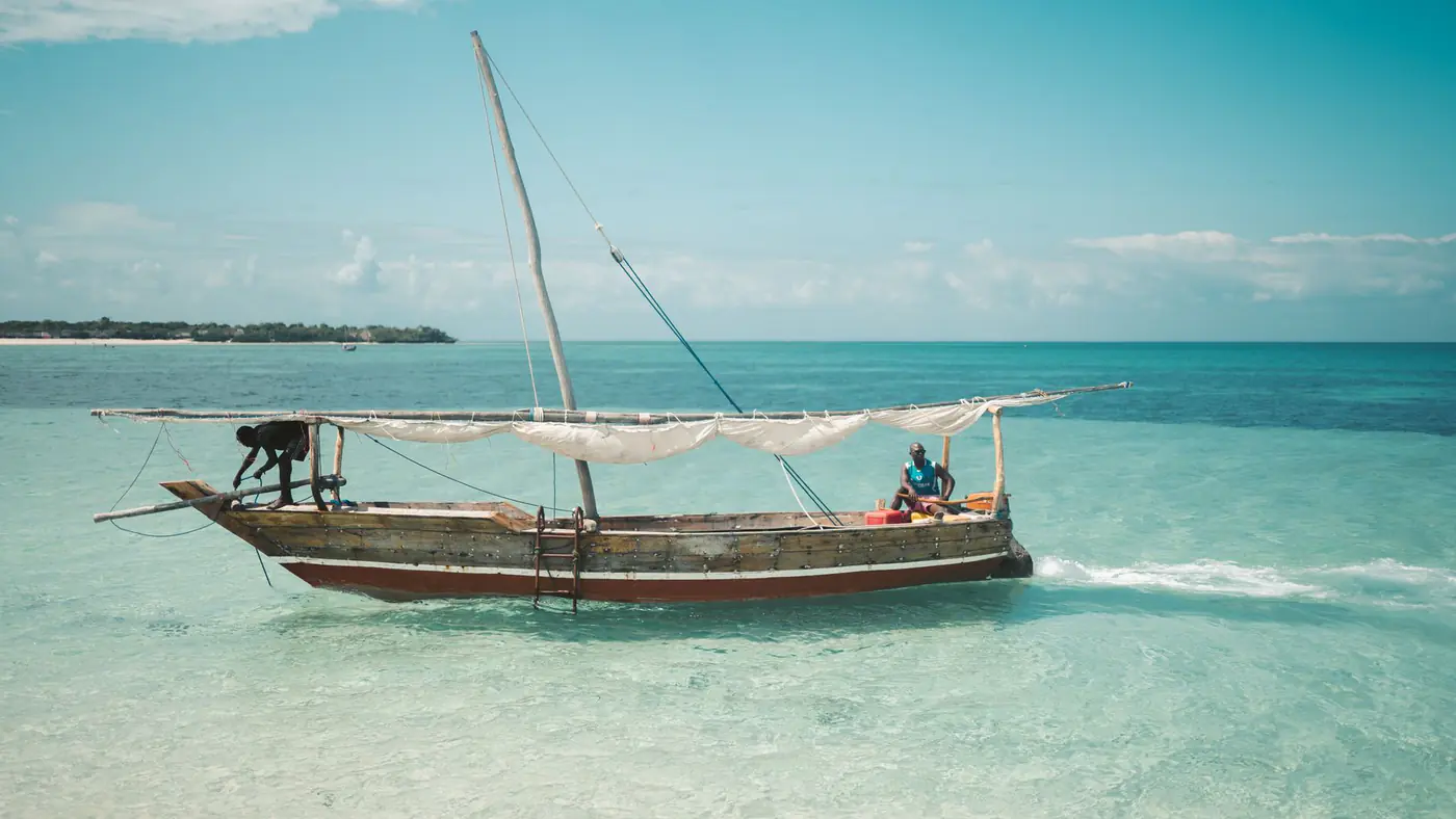 Two local fishermen navigate a traditional dhow on the clear waters of Zanzibar under a bright blue sky.