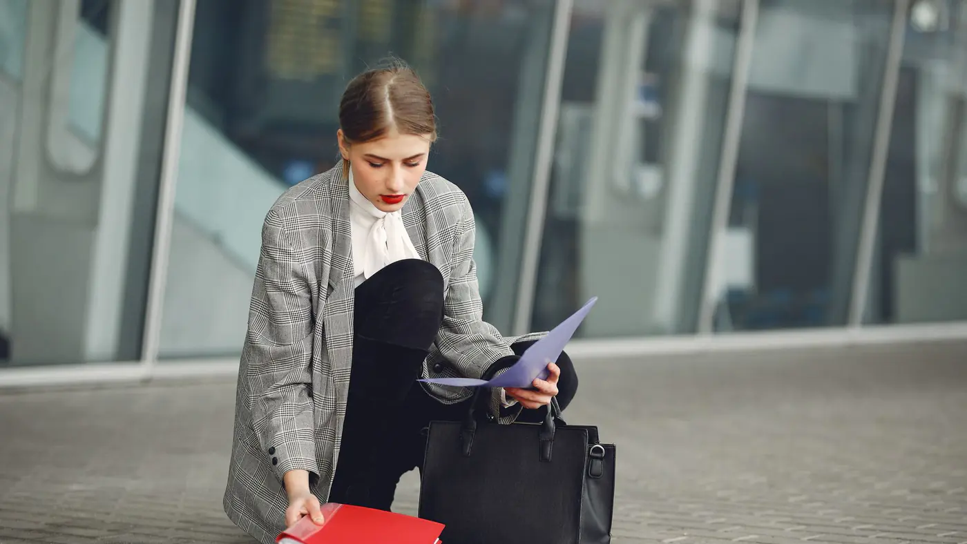 Distracted female manager in trendy wear collecting scattered documents and passport from sidewalk into briefcase near glass wall of modern building